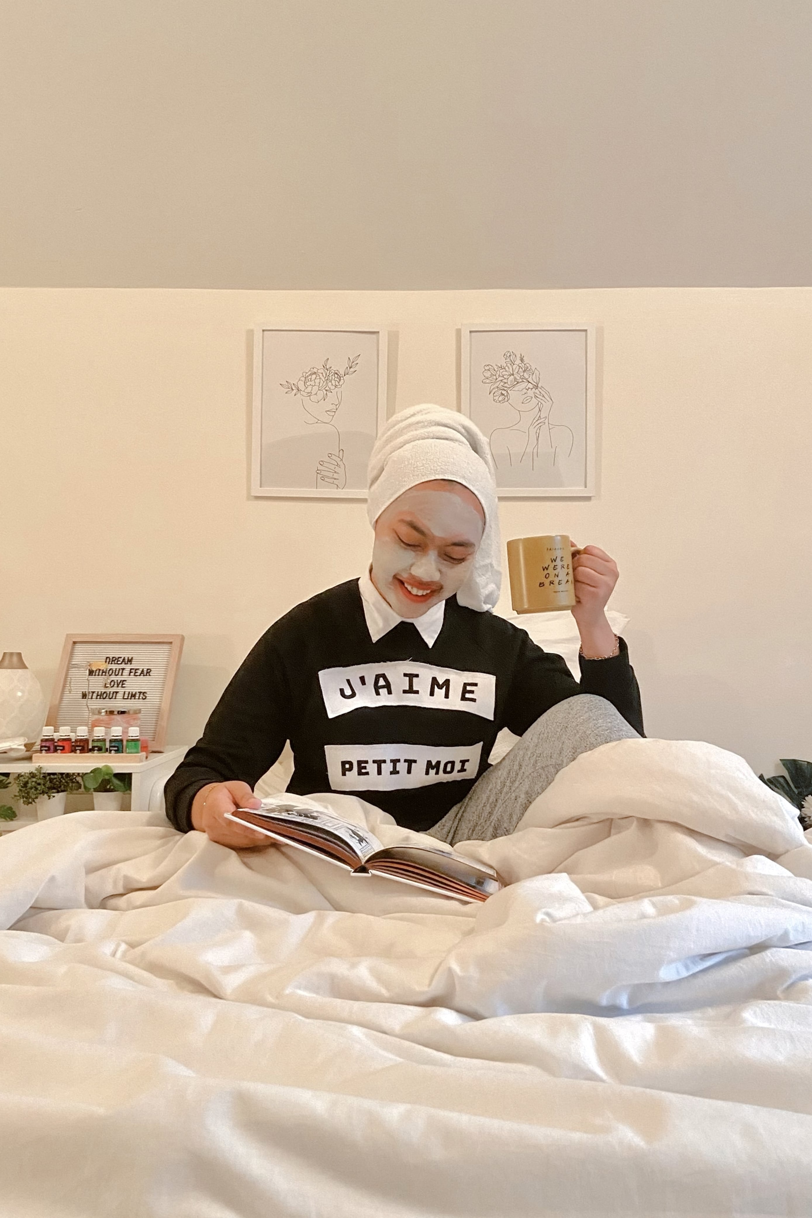 Female model in self-care mode. Wearing high quality black sweater made by petit moi. Sitting down on the bed with a cup of coffee and a book