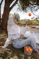 female model catching an orange by the lake wearing a white blouse made by petit moi