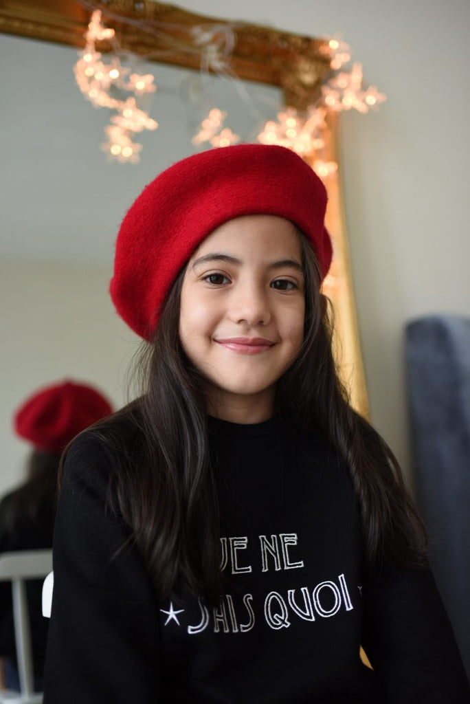 Little girl in high quality black sweater by petit moi. Wearing a red hat and smiling for the camera
